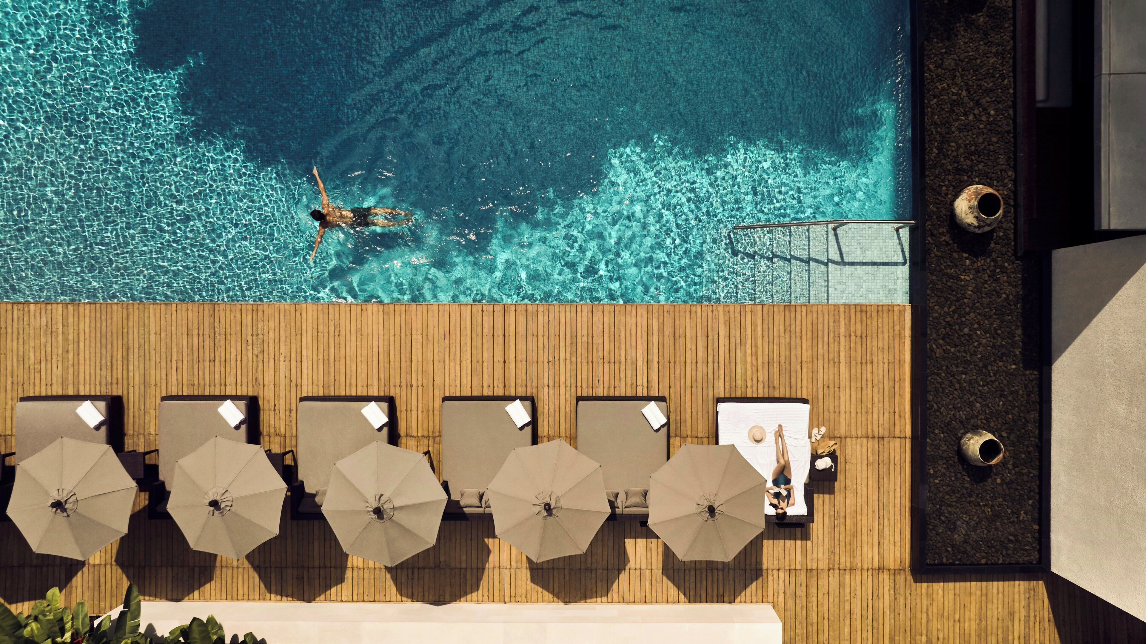 An aerial view of a wooden deck with lounge chairs and umbrellas, overlooking a clear blue pool with a person swimming.