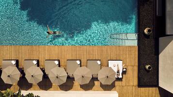 An aerial view of a wooden deck with lounge chairs and umbrellas, overlooking a clear blue pool with a person swimming.