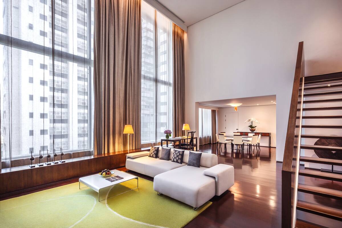 A modern hotel room with a large window, a sofa, a dining area, and a staircase.