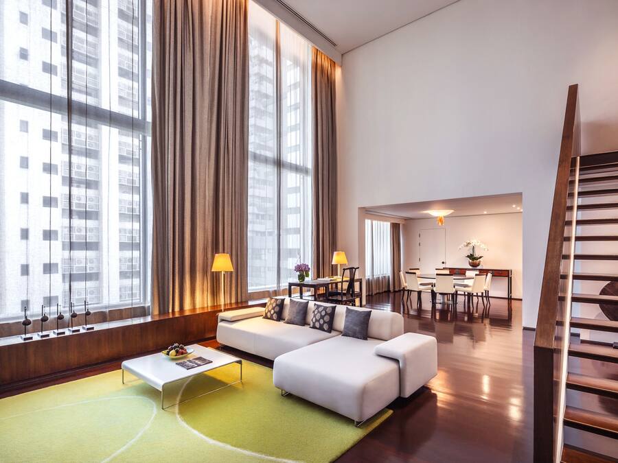 A modern hotel room with a large window, a sofa, a dining area, and a staircase.