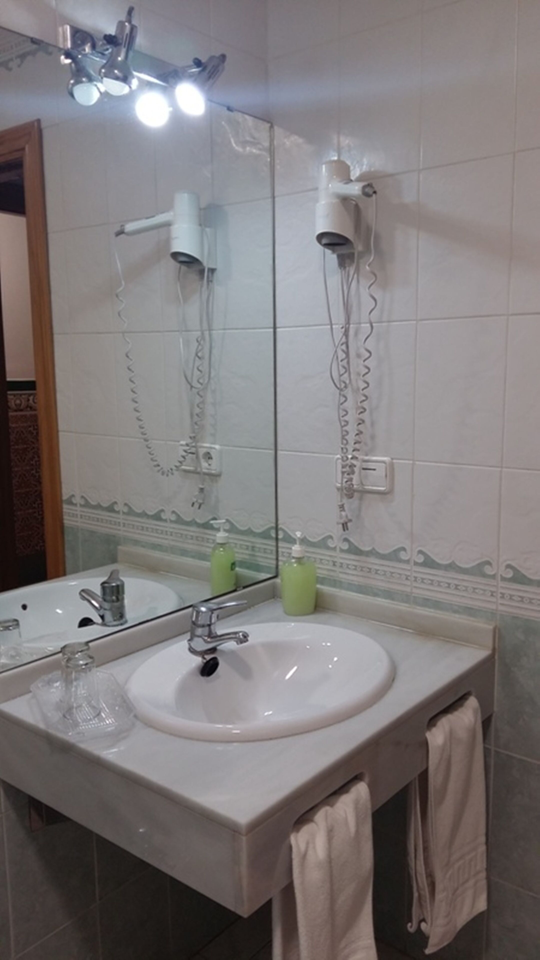Un baño con un espejo grande, dos secadores de pelo fijados en la pared, un lavabo con grifo y dos botellas verdes sobre el mostrador.