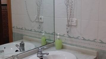 Un baño con un espejo grande, dos secadores de pelo fijados en la pared, un lavabo con grifo y dos botellas verdes sobre el mostrador.