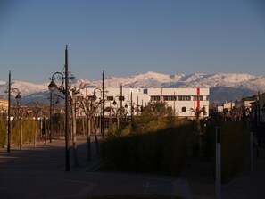 A hotel with a modern design, featuring a prominent red stripe on the facade, situated in a town with a backdrop of snow-capped mountains. - Hotel Corona de Atarfe (Atarfe)