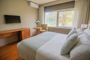 A hotel room with a bed, a desk, a chair, a TV, and a window with a view of trees. - Wyndham Garden Campana (Campana)