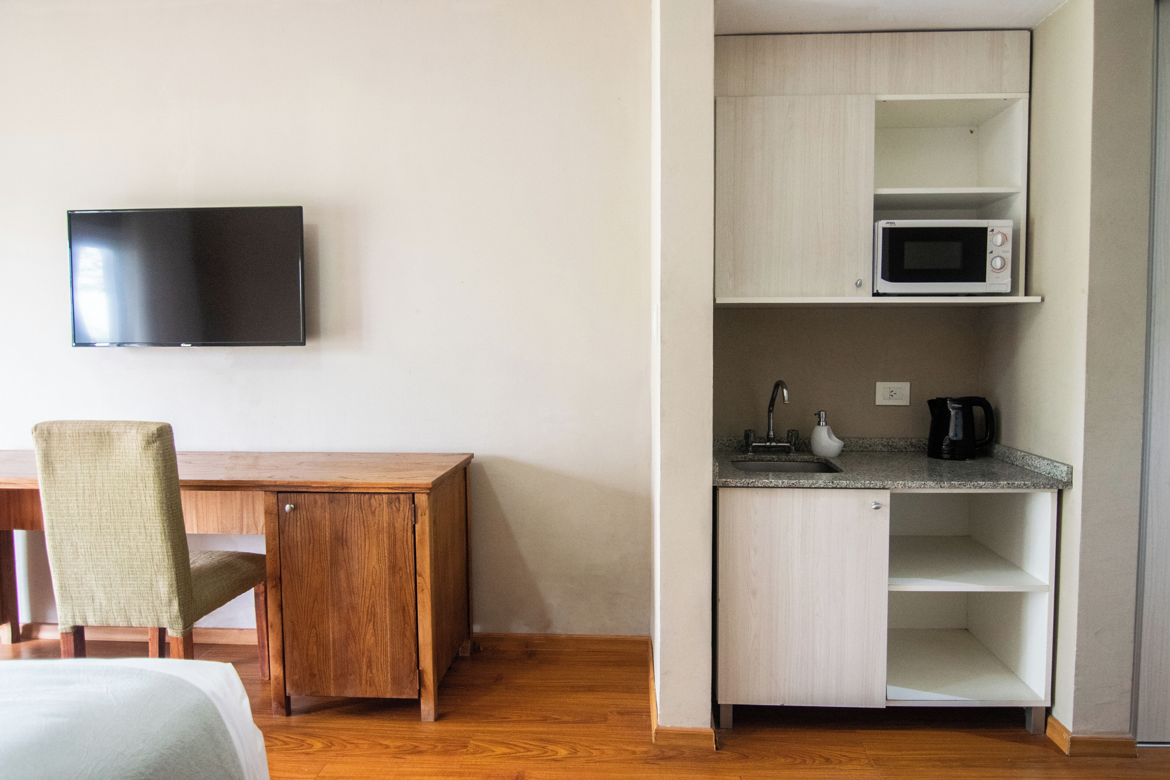 A compact room with a wooden desk, a television mounted on the wall, a microwave on a shelf, and a chair.