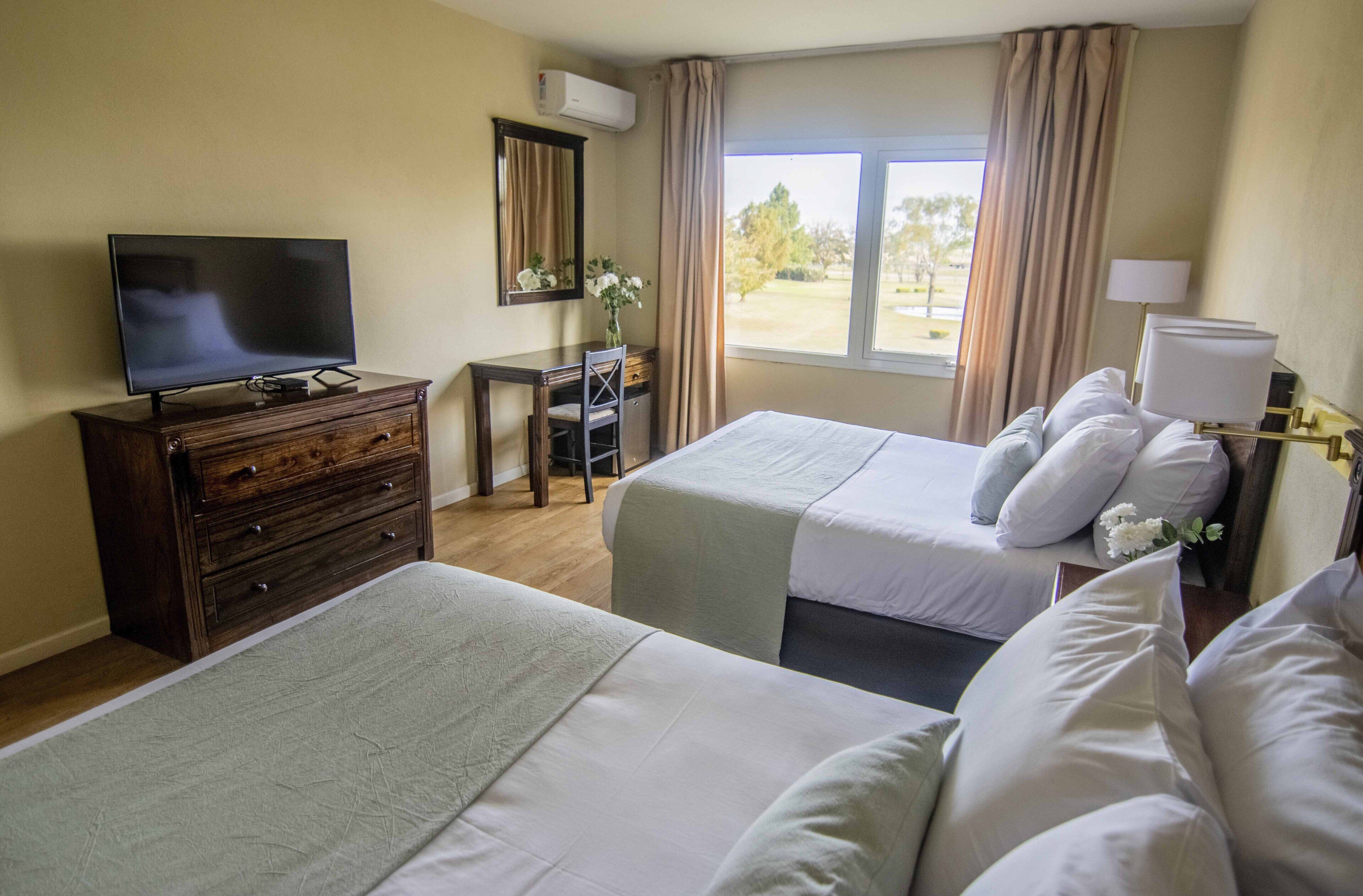 Ein Hotelzimmer mit zwei Betten, einem Fernseher, einem Schreibtisch, einem Stuhl, einer Kommode, einem Fenster mit Vorhängen und einer Lampe.