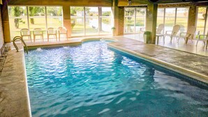 Piscina interior con agua cristalina, rodeada de sillas y grandes ventanales que ofrecen vistas a un jardín ajardinado.