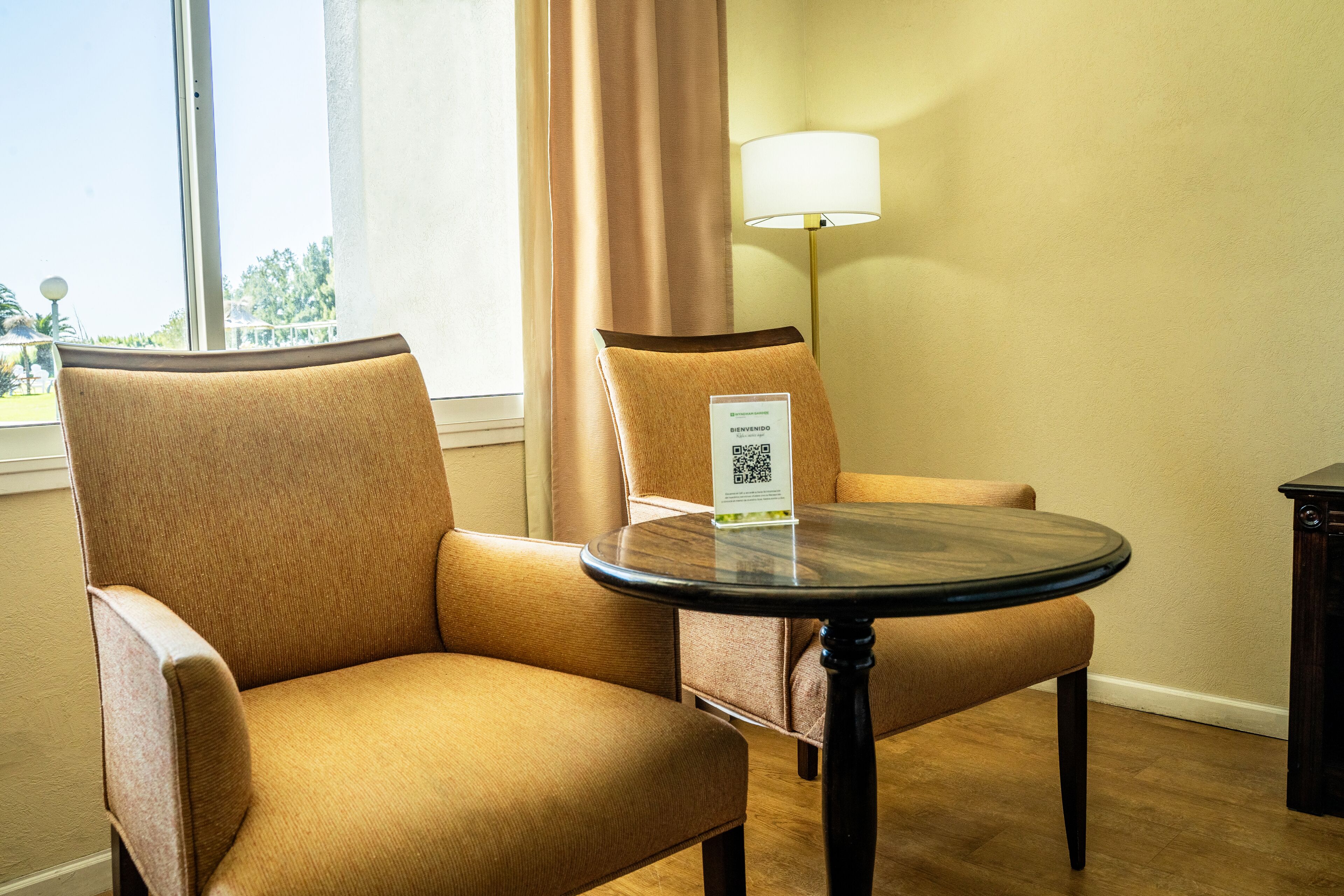 Habitación de hotel con dos sillones color beige, una mesa redonda, una lámpara y un cartel con un código QR.