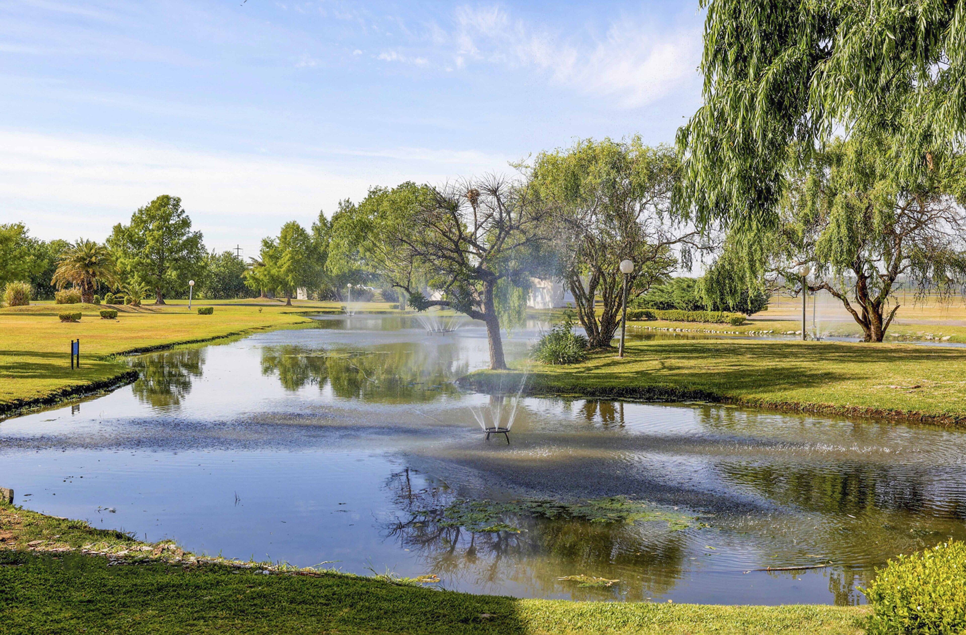 Un étang paisible avec une fontaine au centre, entouré de pelouses et d’arbres bien entretenus.