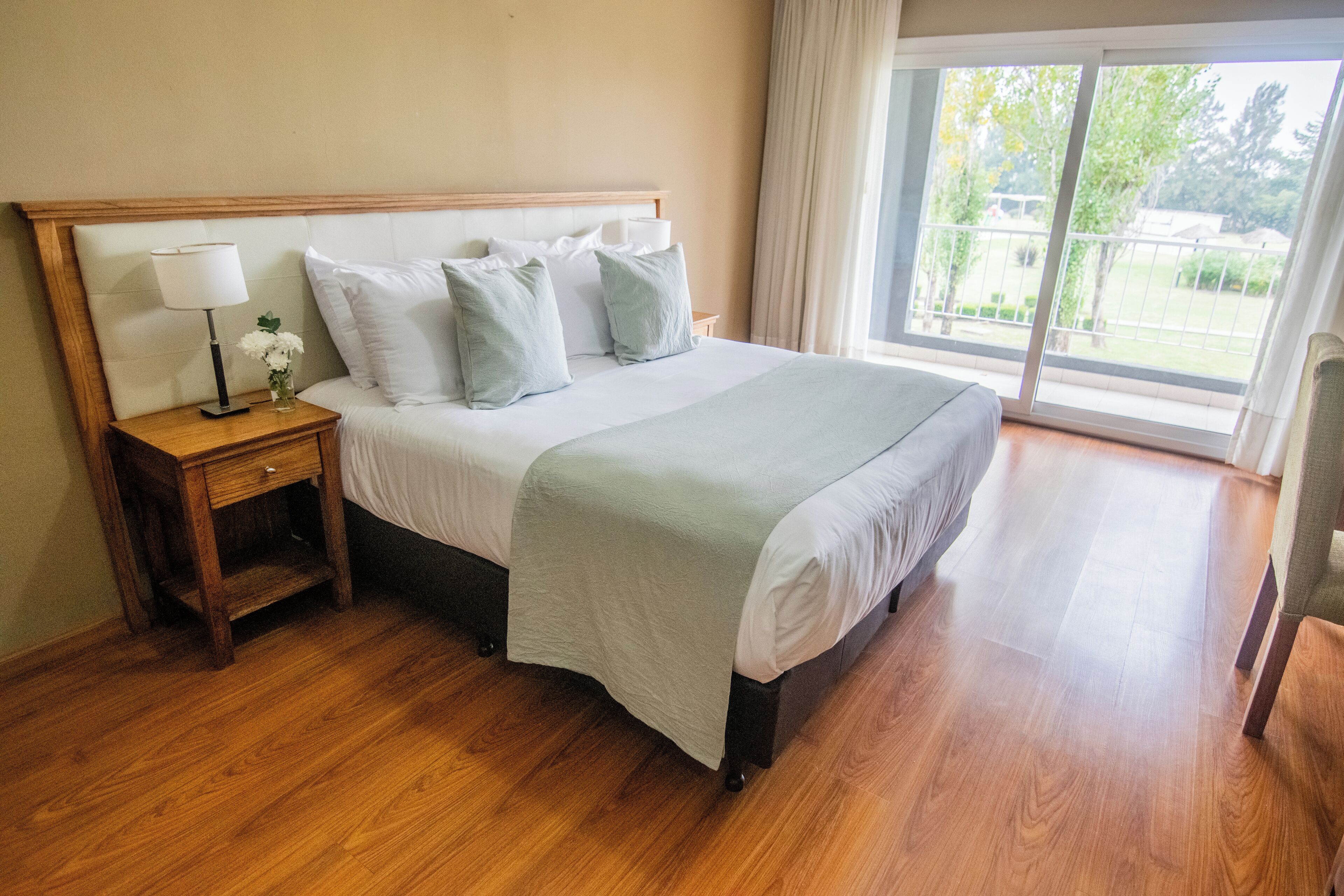 A neatly made bed with a wooden headboard, a bedside table with a lamp and a vase of flowers, and a large window with a view of greenery outside.