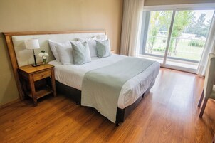 Una cama bien hecha con cabecera de madera, una mesita de noche con lámpara y un florero, y un ventanal con vistas a zonas verdes.