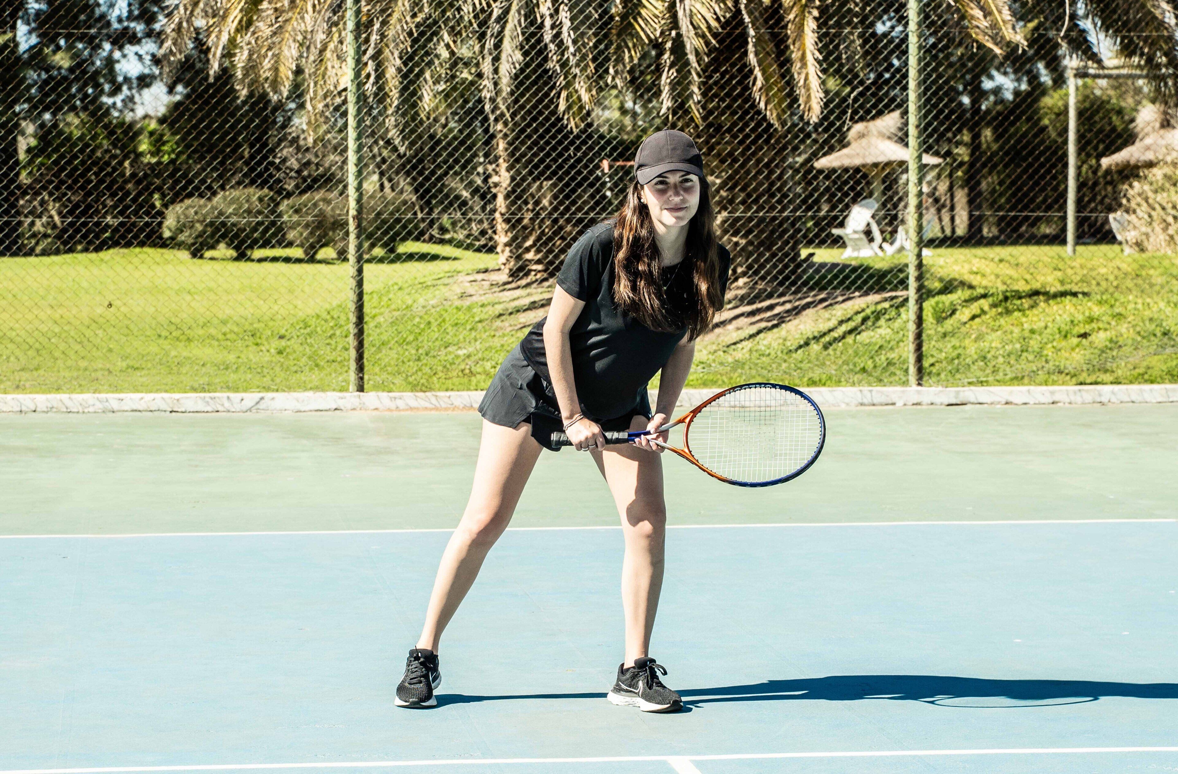 A tennis court with a player in a black outfit holding a racket, a chain-link fence, and trees in the background.