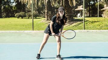 A tennis court with a player in a black outfit holding a racket, a chain-link fence, and trees in the background.