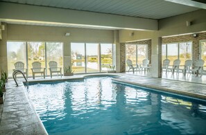 An indoor swimming pool with clear glass walls, surrounded by white chairs, and a view of a landscaped area outside. - Wyndham Garden Campana (Campana)