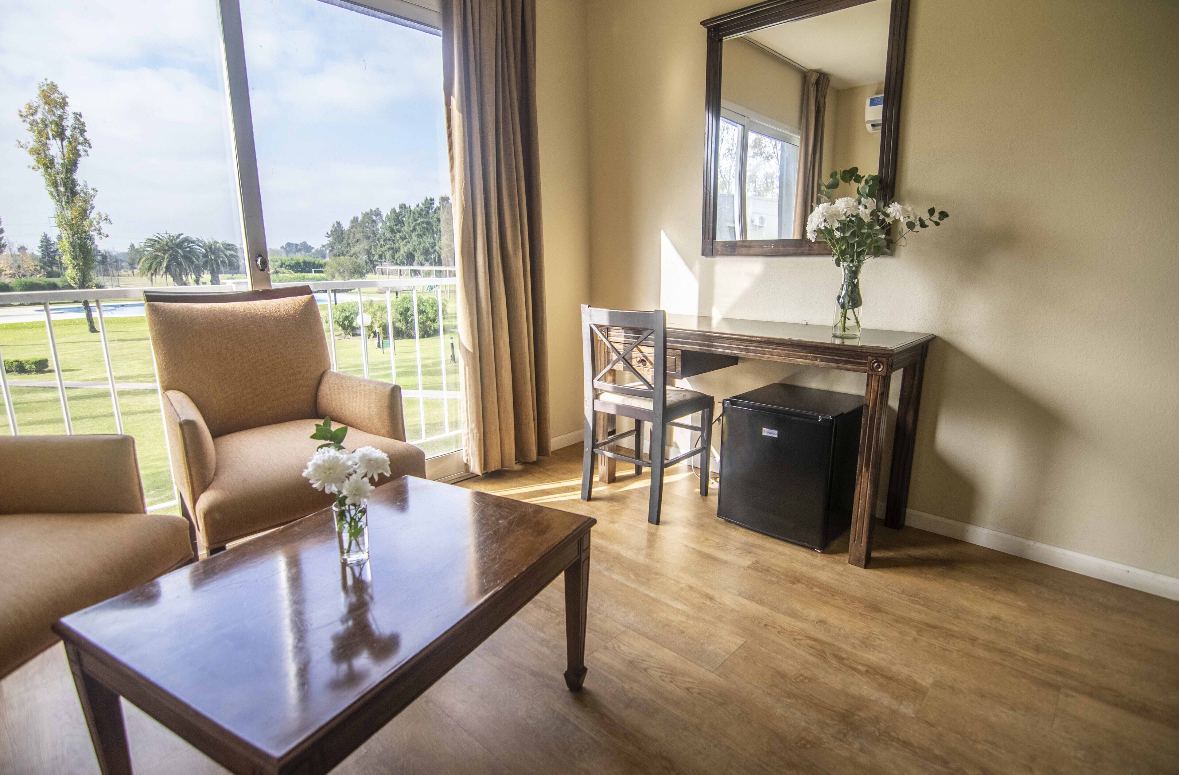 Habitación con vista a un campo de golf, un escritorio, una silla, un refrigerador pequeño y una mesa de centro con un jarrón de flores.