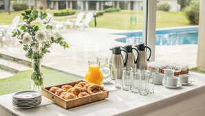 Desayuno con croissants, zumo de naranja, cafeteras y tazas de té sobre una mesa cerca de una piscina.