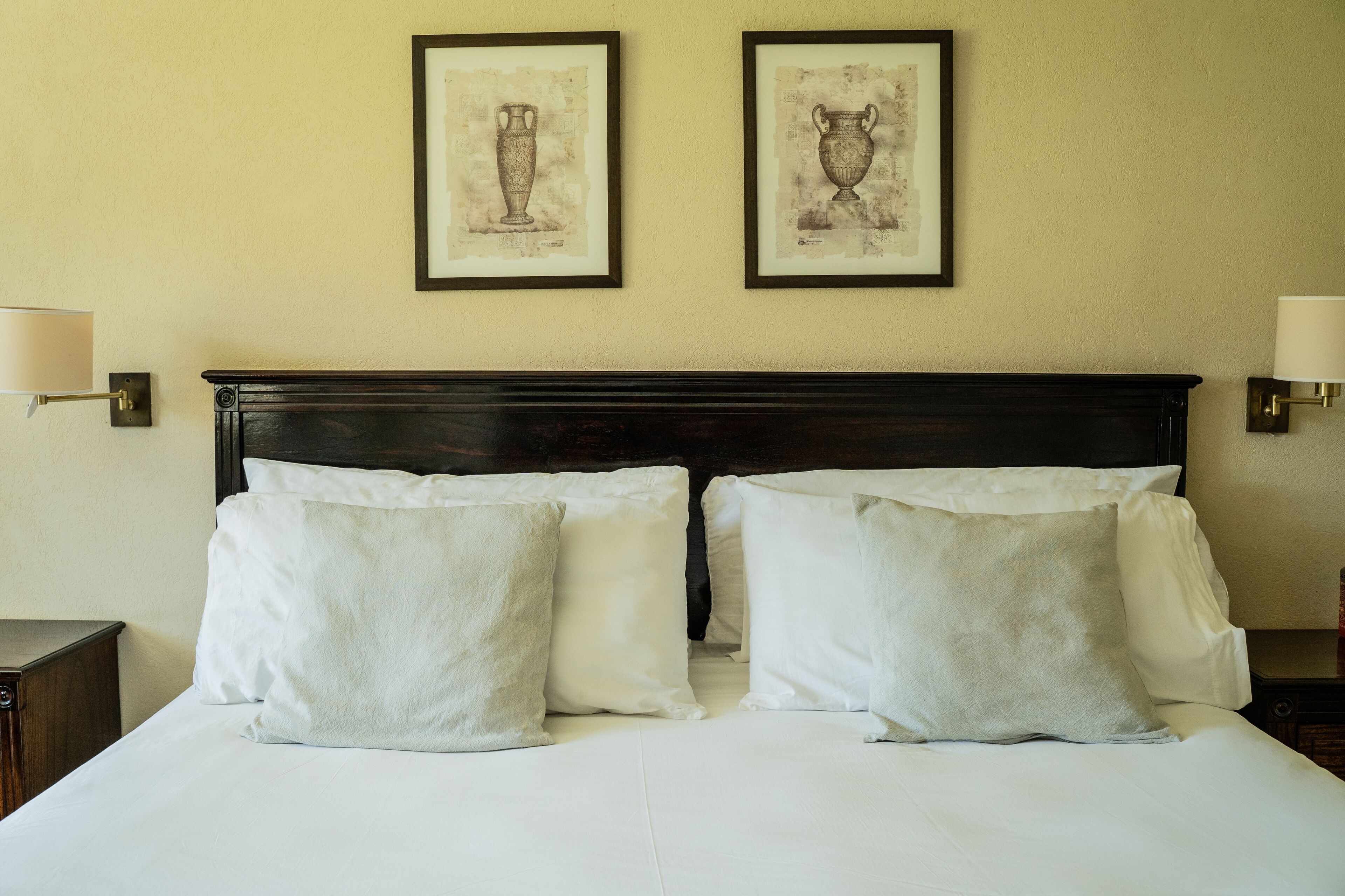 A neatly made bed with white linens and pillows, a dark wooden headboard, and two framed artworks on the wall.