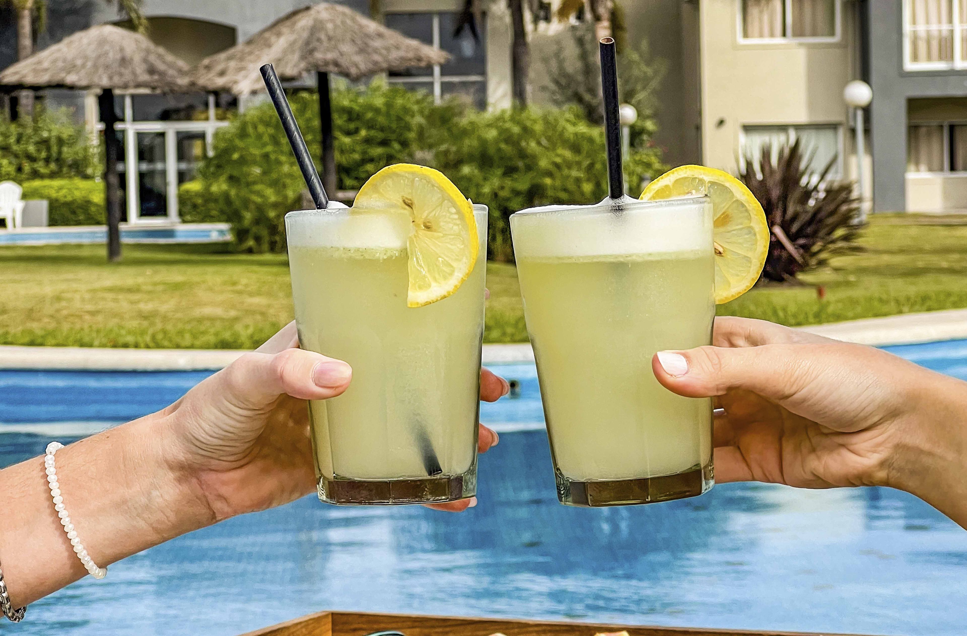 Duas mãos segurando copos com bebidas e fatias de limão, perto de uma piscina com guarda-sóis e um edifício ao fundo.