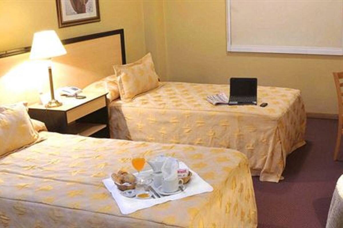 Ein Hotelzimmer mit zwei Betten, einem Nachttisch mit Lampe, einem Laptop auf einem Bett und einem Frühstückswagen auf dem anderen.