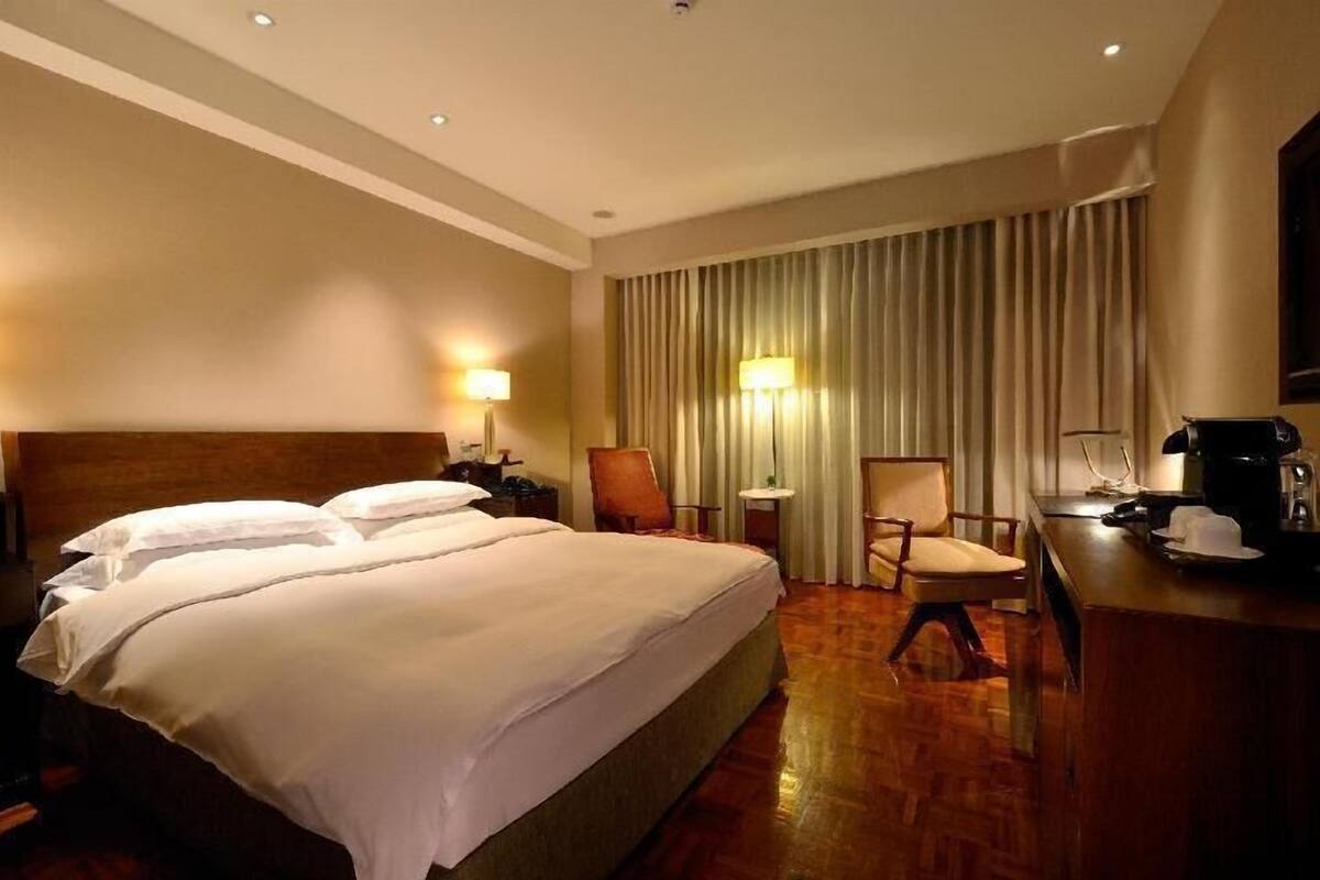 A hotel room with a large bed, two bedside tables, a desk with a chair, a lamp, and a window with curtains.