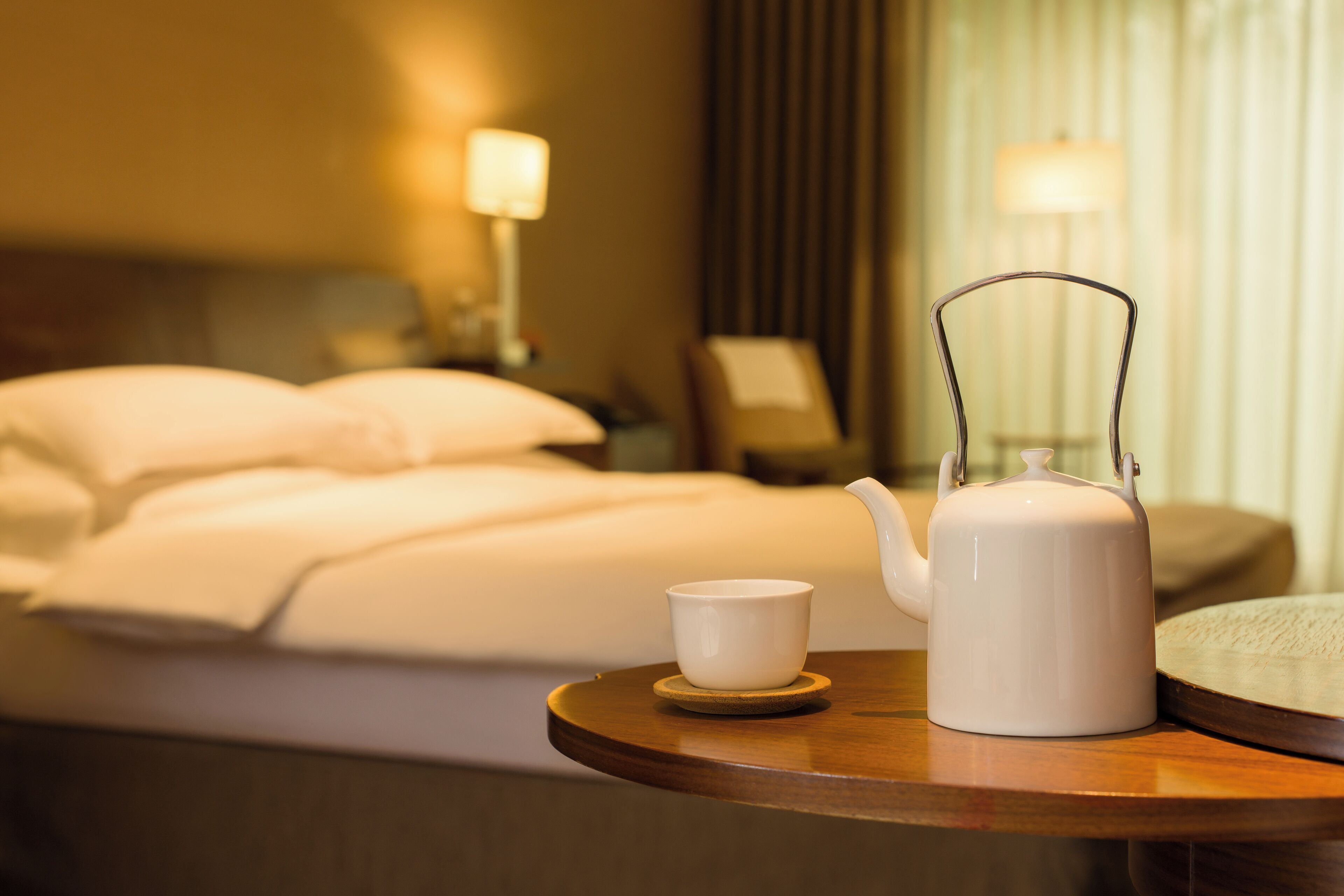 Habitación de hotel con cama, mesita de noche con tetera y taza, y una lámpara.