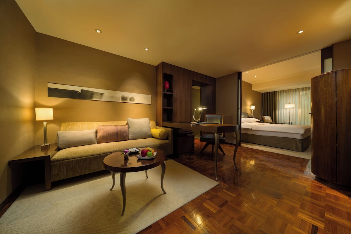 A hotel room with a sofa, a small table with fruit, a desk with a lamp, and a bed.