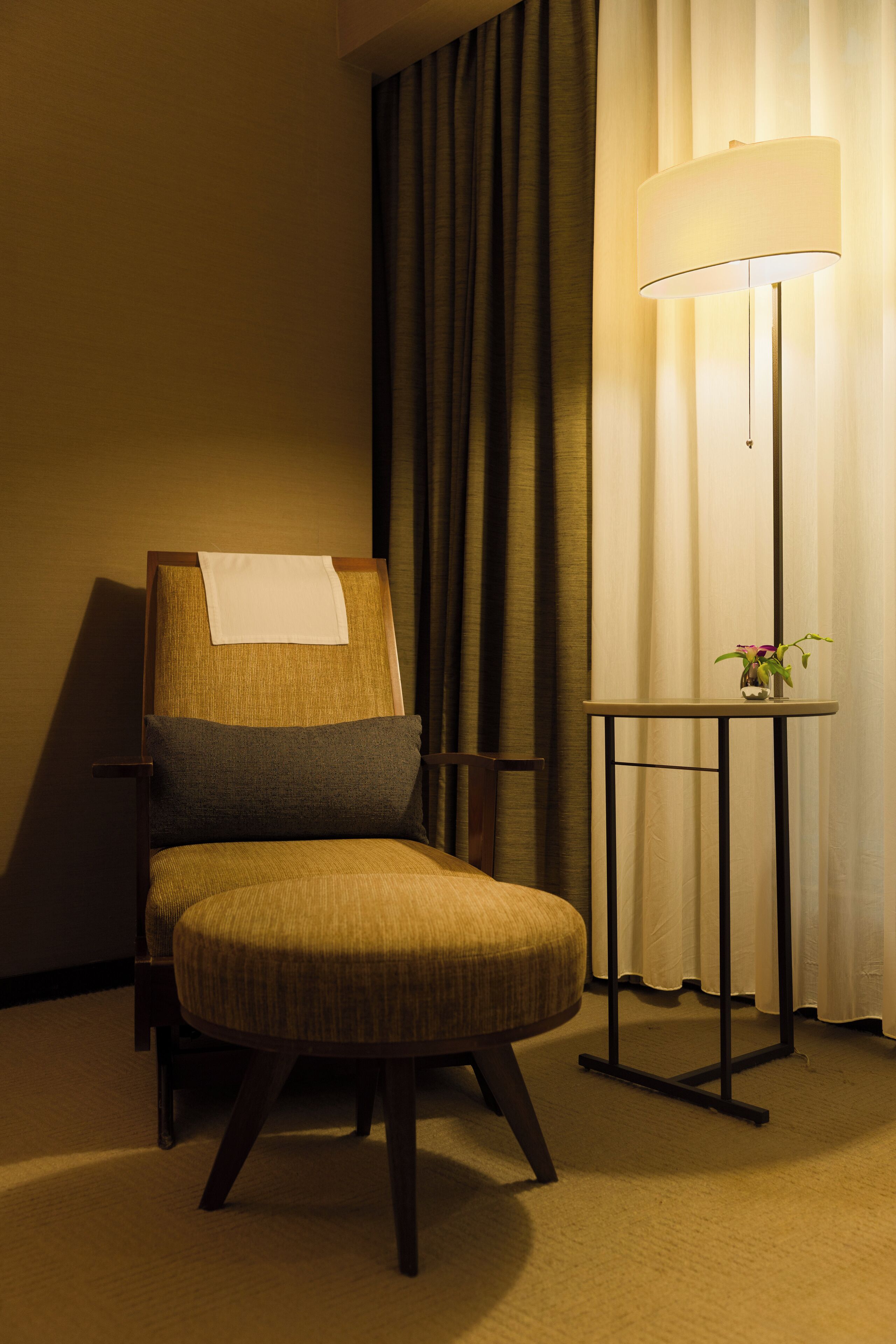 A hotel room with a single armchair, a small table with a lamp, and a curtain.