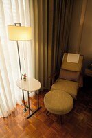Een kamer met een houten vloer, een ronde tafel met een lamp, een fauteuil en een raam met gordijnen.