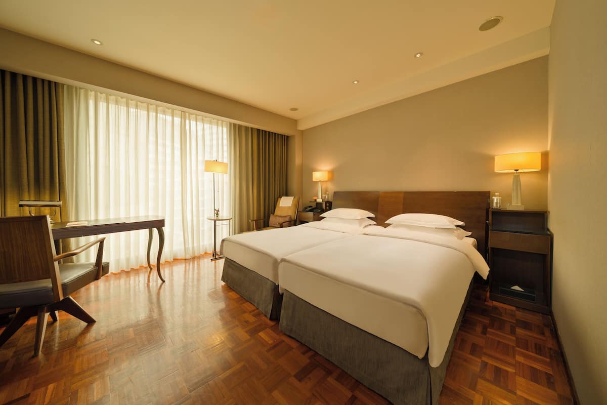 A hotel room with two beds, a desk, a chair, and a lamp.