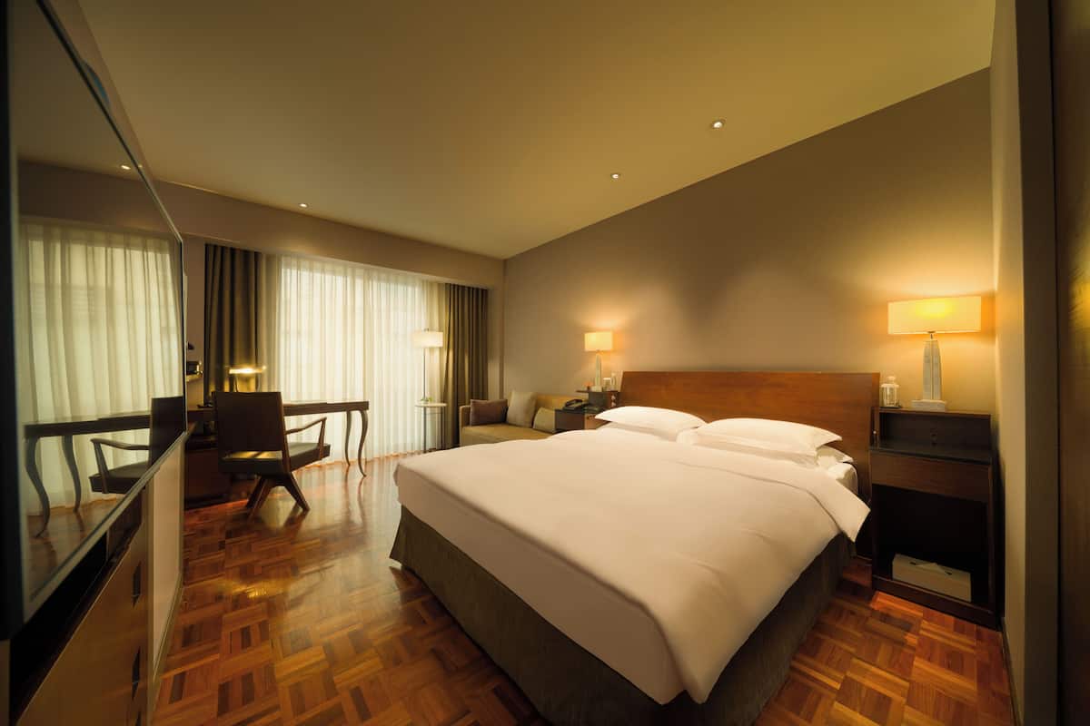 A hotel room with a large bed, a desk with a chair, a nightstand with a lamp, and a mirror.