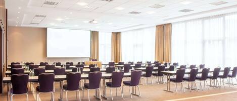 Un salón de conferencias con filas de mesas y sillas, una pantalla de proyector y ventanas amplias con cortinas.