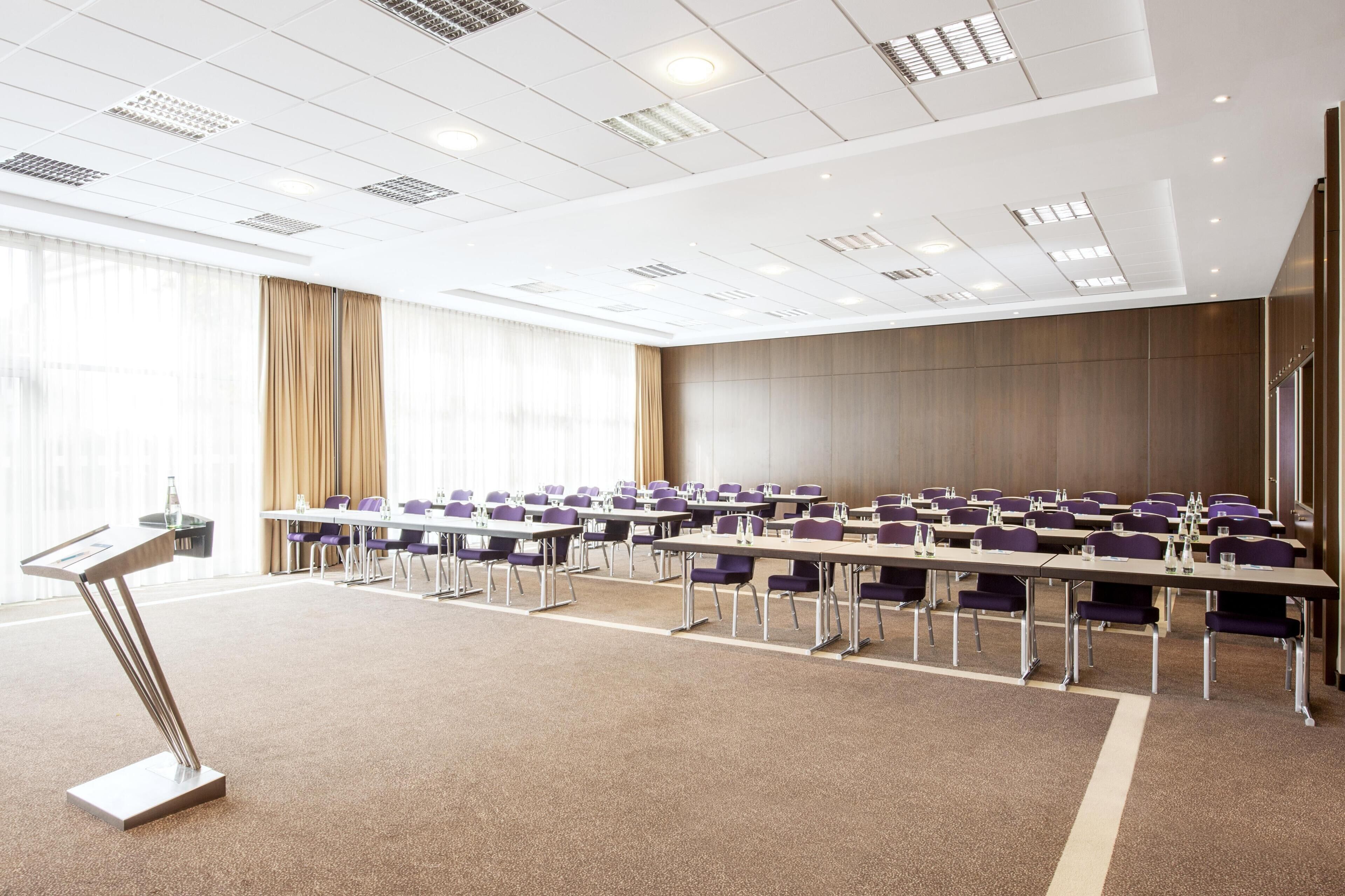 A conference room with rows of tables and chairs, a podium, and large windows with curtains.