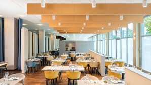 A modern dining area with multiple tables and chairs, large windows, and a high ceiling with recessed lighting.