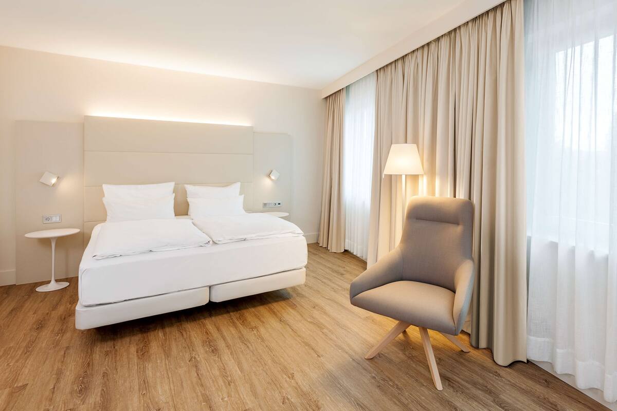 A modern hotel room with a large bed, a bedside table, a chair, and a window with curtains.