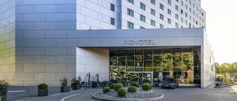 Un hotel moderno con fachada de vidrio, entrada despejada y un área ajardinada con arbustos bien cuidados.