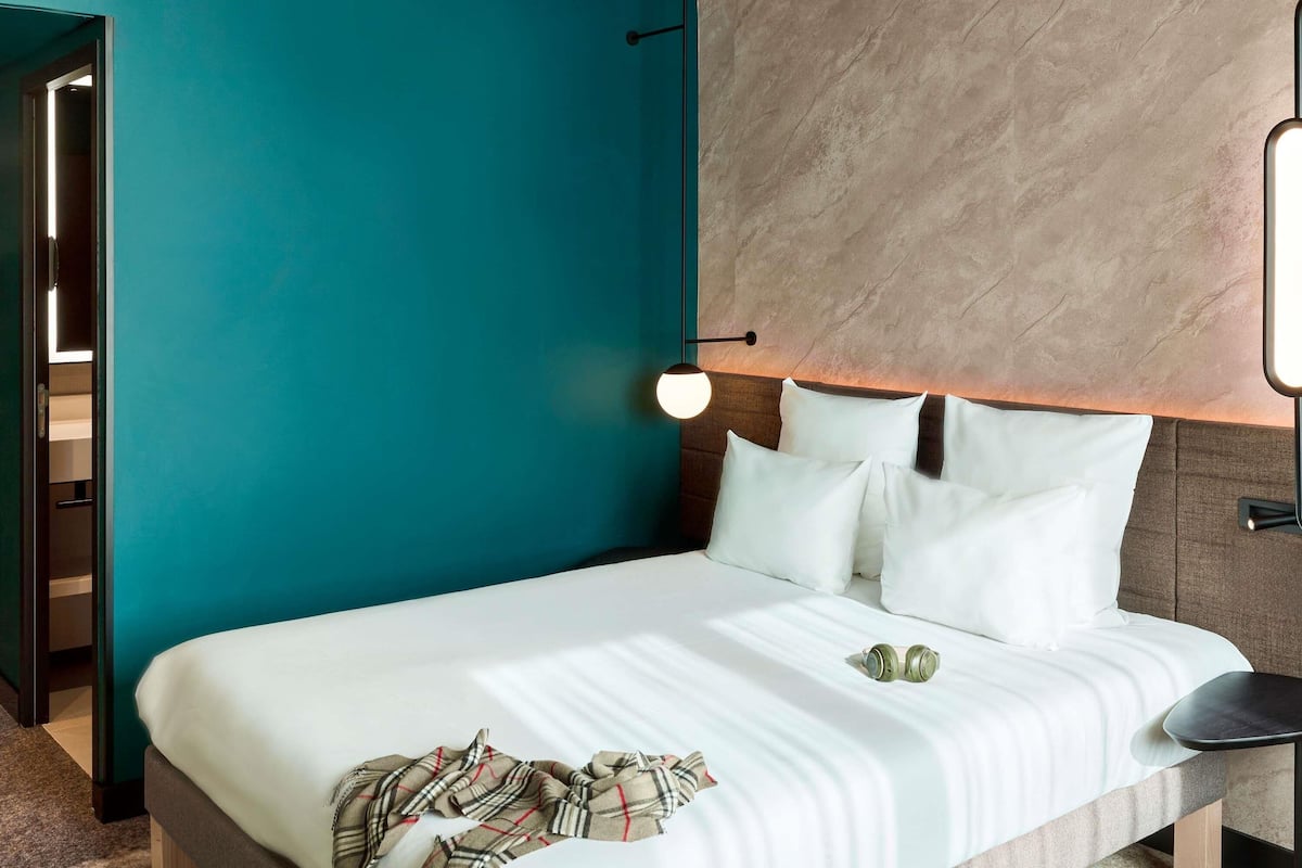 A modern hotel room with teal walls, a wooden headboard, a bed with white bedding, a bedside table, and a wall-mounted light fixture.