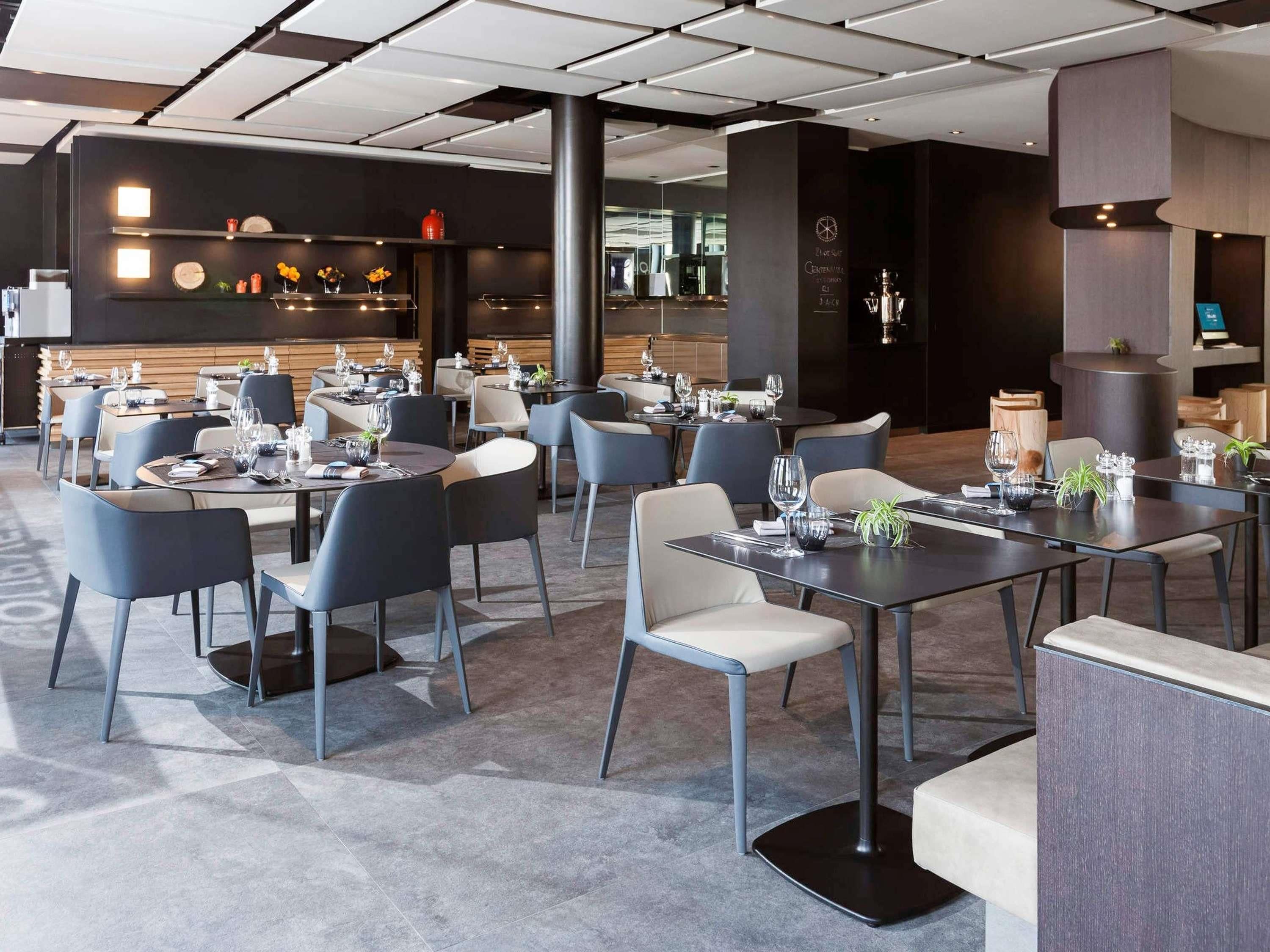 A modern restaurant with round tables, grey chairs, and a bar area in the background.