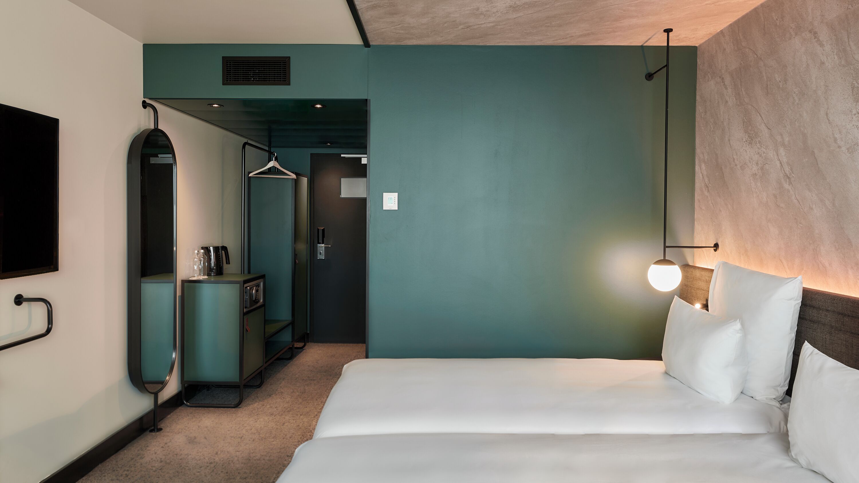 A hotel room with a bed, a wardrobe, a mirror, a desk, and a hanging light fixture.
