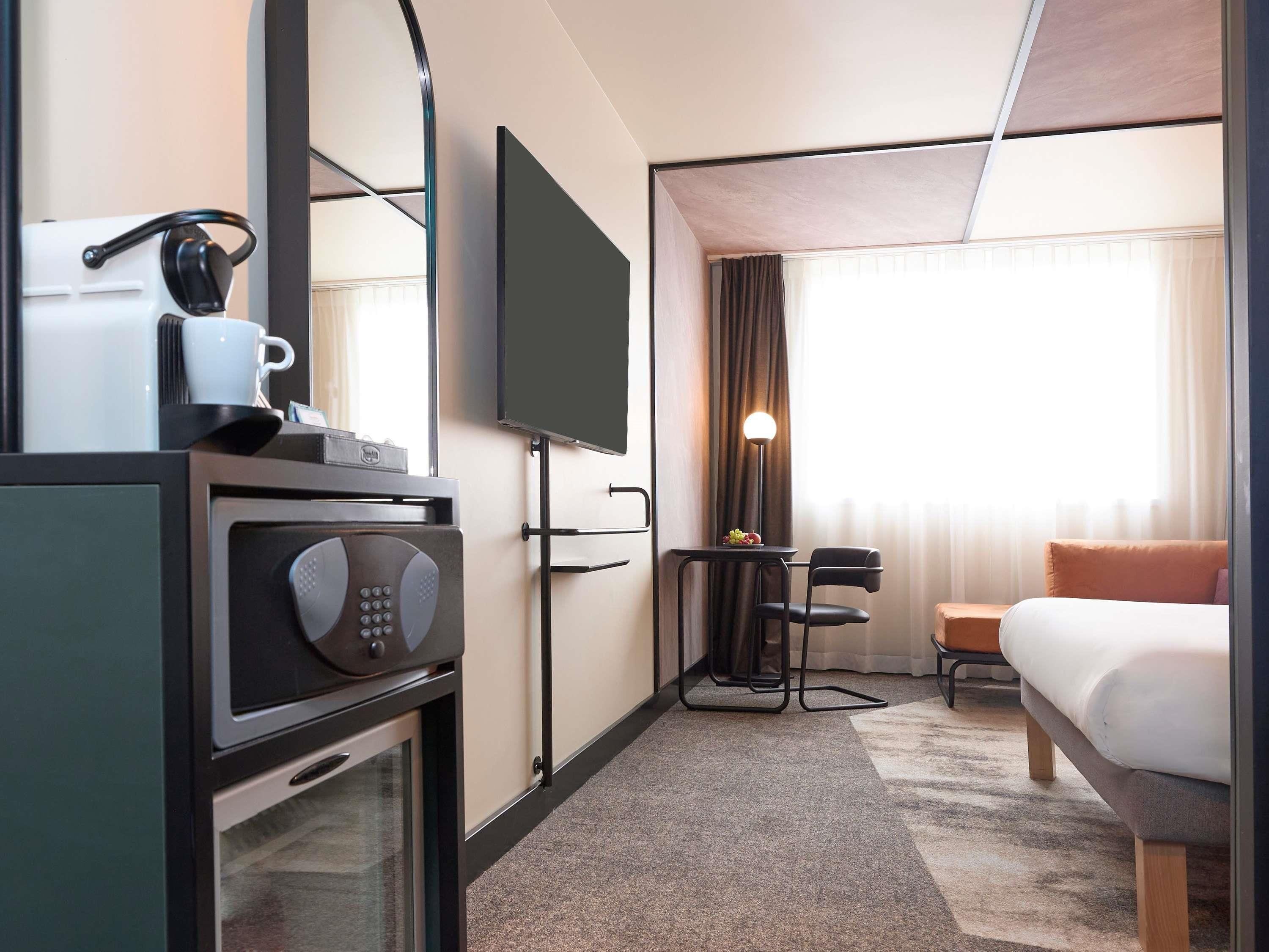A hotel room with a bed, a desk with a chair, a television, a coffee machine, and a window with curtains.