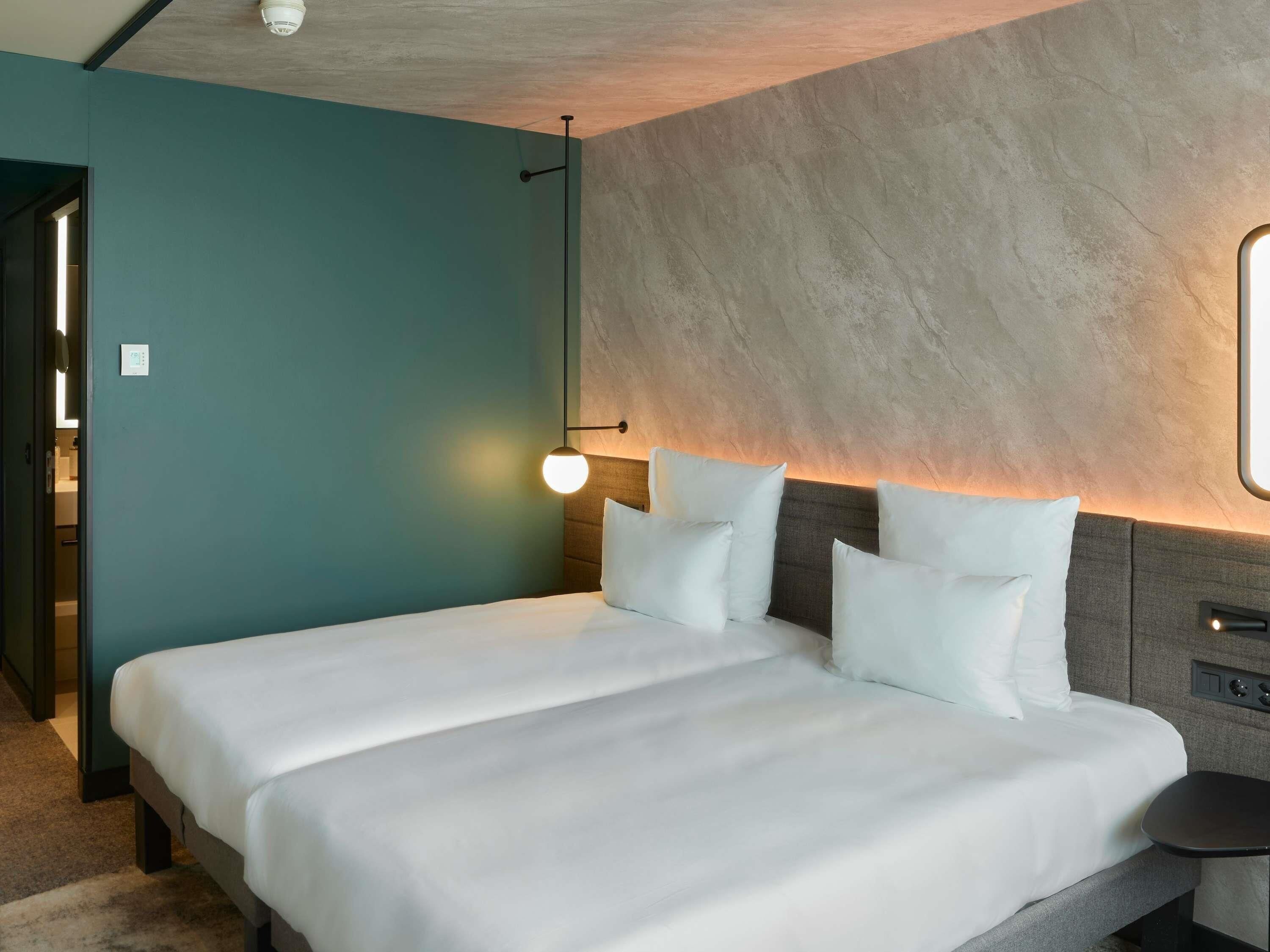 Una habitación de hotel con cama, mesita de noche, lámpara en la pared y puerta que da a un baño.