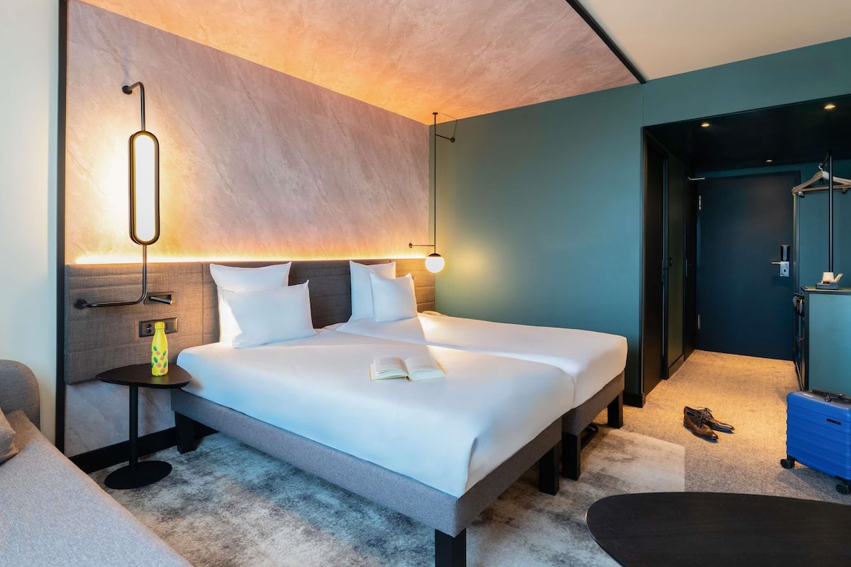 Habitación de hotel moderna con una cama grande, mesitas de noche, una lámpara de lectura y vistas al pasillo.