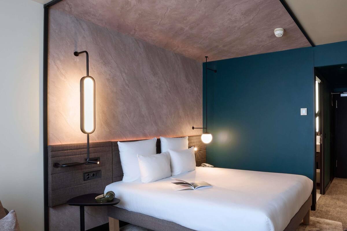 Quarto de hotel moderno com cama grande, mesas de cabeceira, luminária na parede e luz no teto.