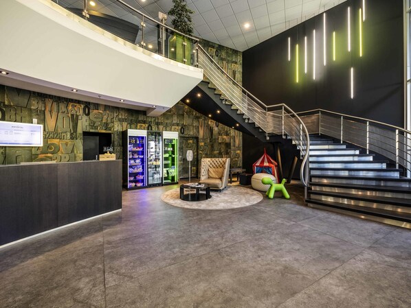 A modern lobby with a reception desk, vending machines, a staircase, and a seating area with a round table and chairs.