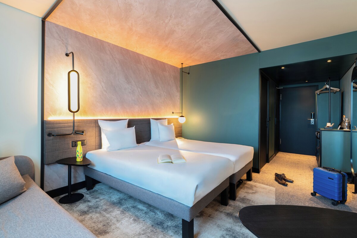 A modern hotel room with a large bed, bedside tables, a small table with a bottle, a suitcase, and a mirror.