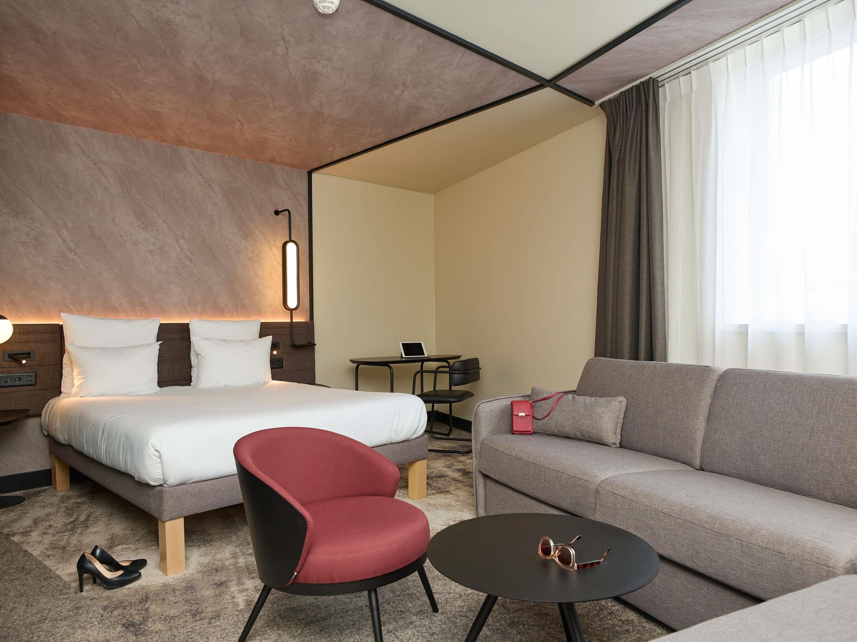 A hotel room with a bed, a red chair, a grey sofa, a small round table, and a desk with a chair.