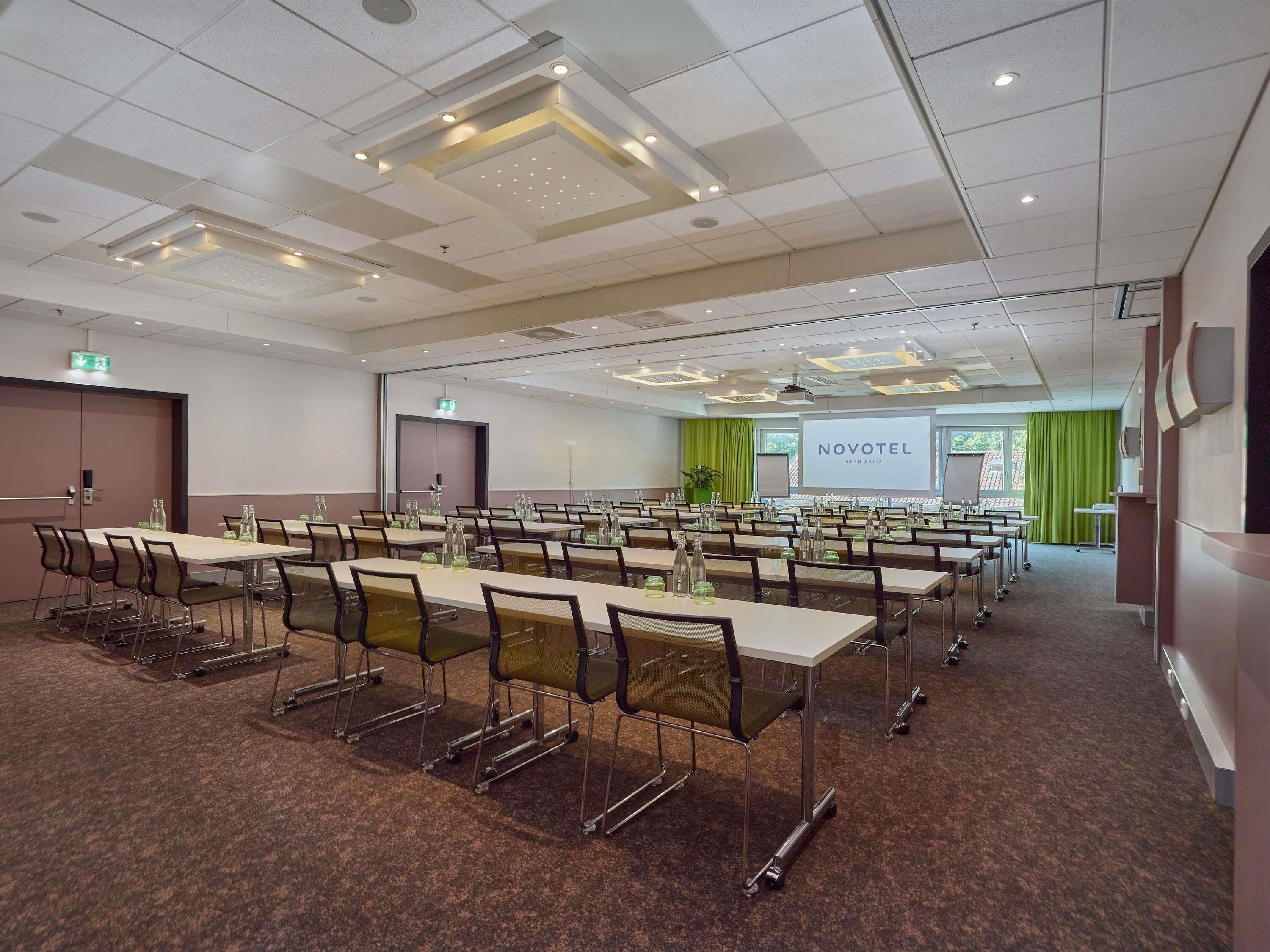 A conference room with multiple tables and chairs, a projector screen, and a green curtain.