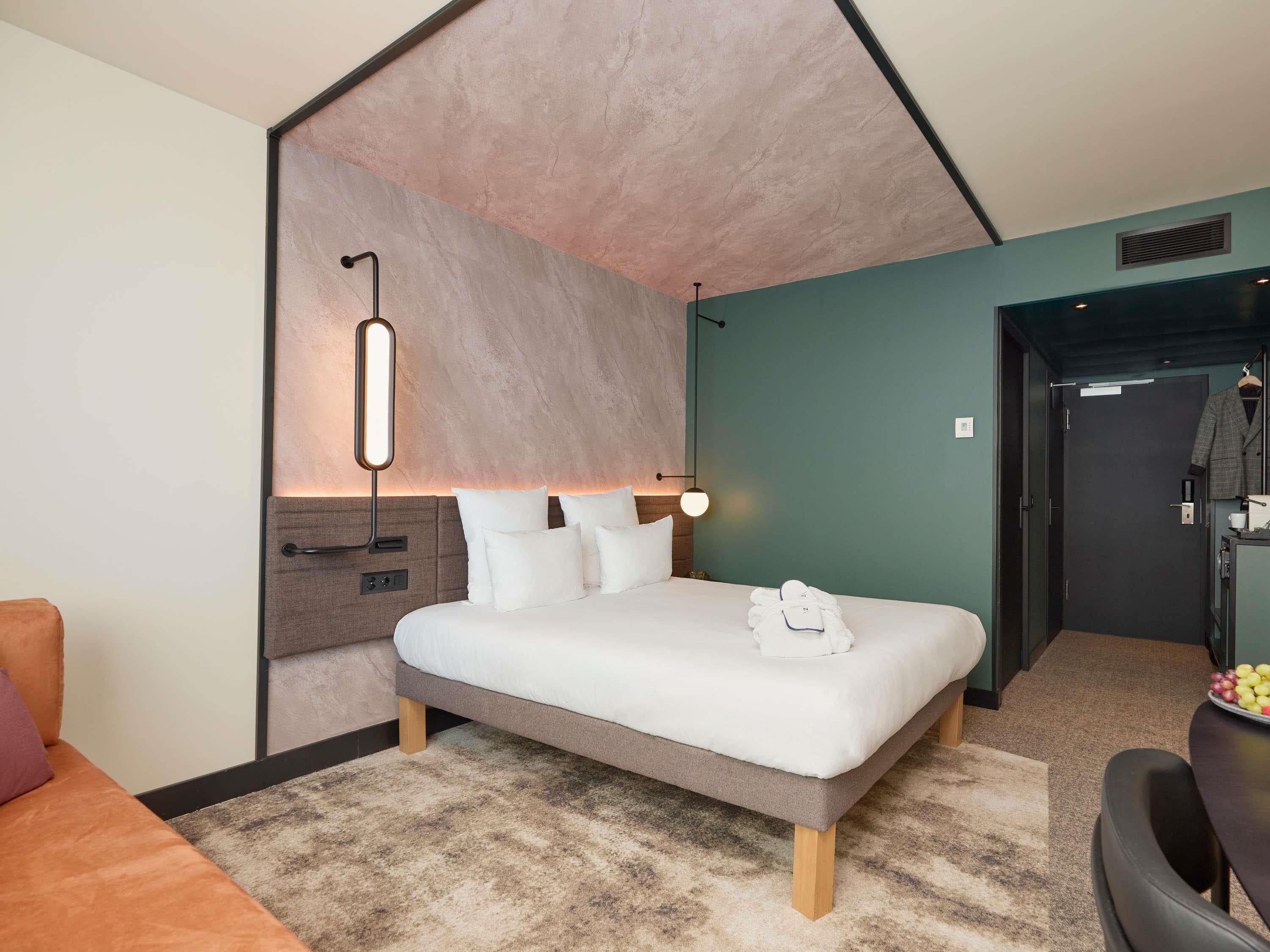 Una habitación de hotel moderna con cama, mesita de noche, lámpara y vistas al pasillo.