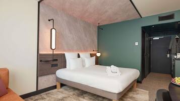 Una habitación de hotel moderna con cama, mesita de noche, lámpara y vistas al pasillo.