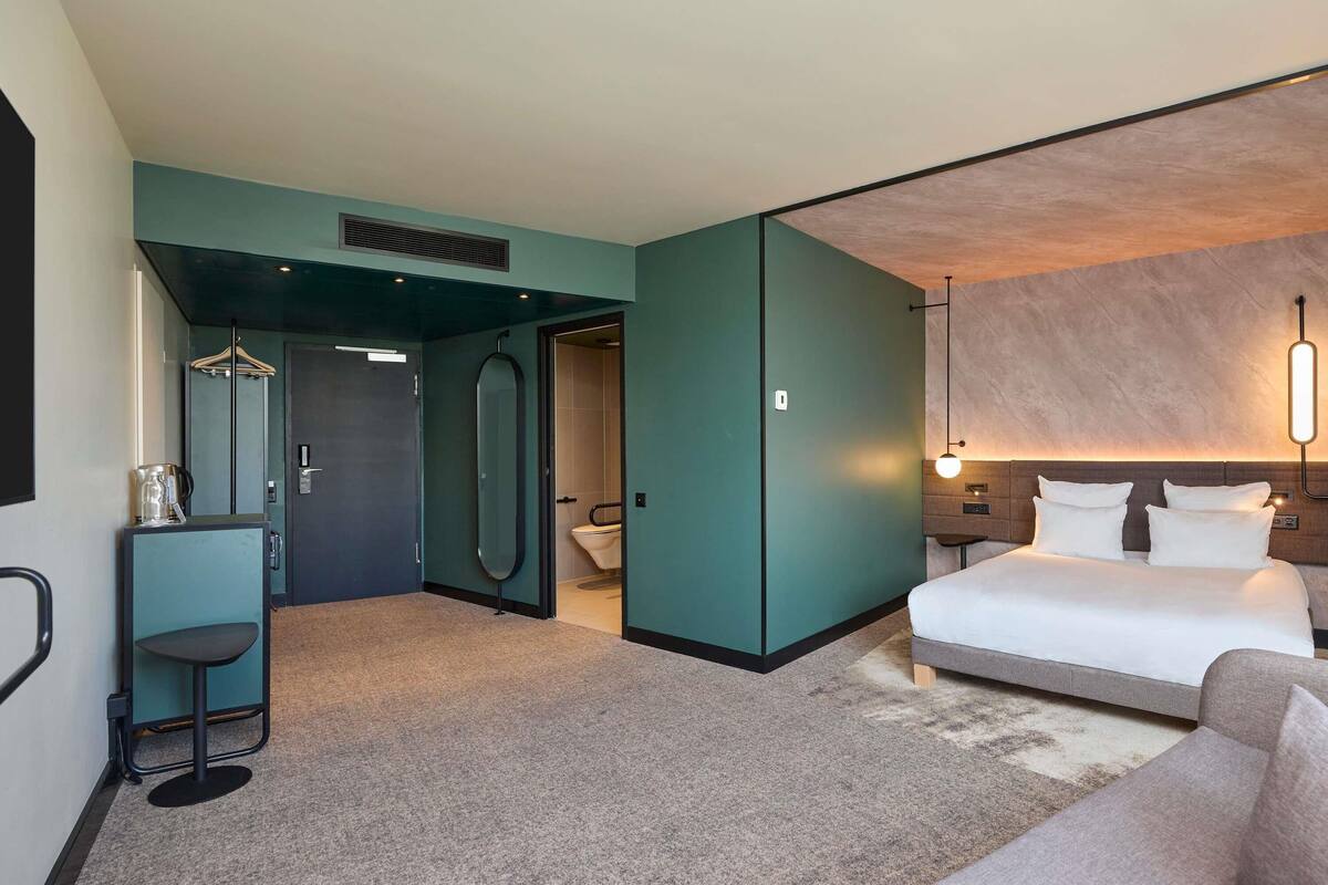Quarto de hotel moderno com cama, área de estar, espelho e uma pequena mesa.