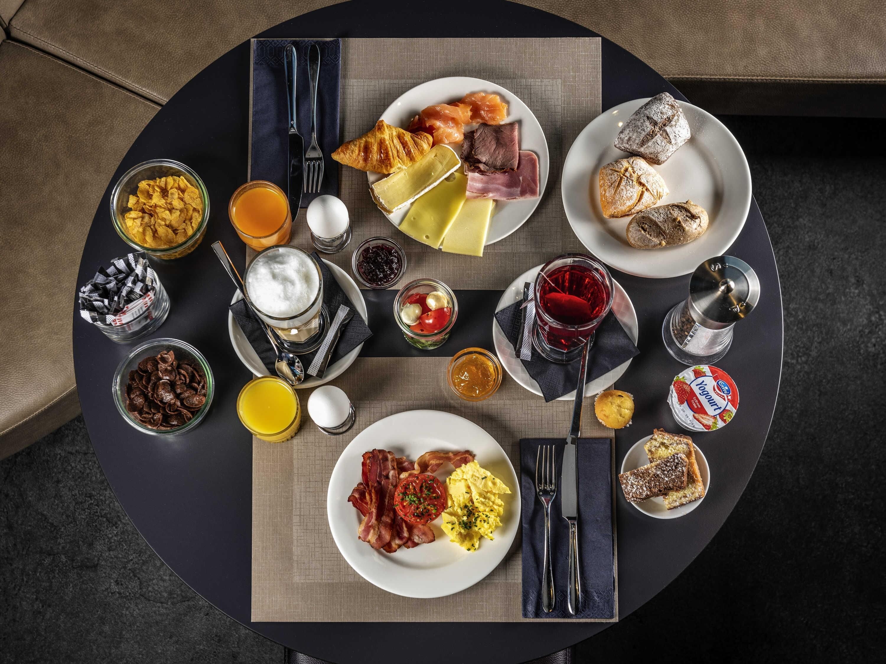 Une table ronde dressée pour le petit-déjeuner, avec des assiettes contenant des œufs, du bacon, des toasts et des viennoiseries.